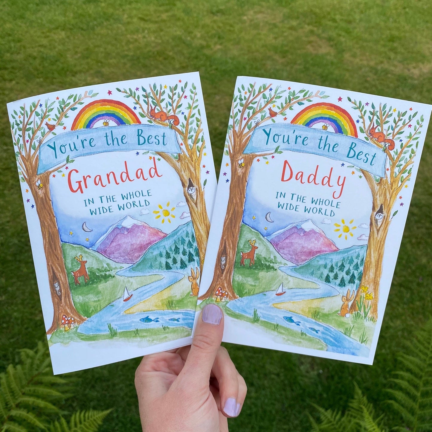 hand holding 2 cards that feature watercolour woodland scenes with mountains in the background. one card says 'you're the best grandad in the whole wide world' and the other 'you're the best daddyin the whole wide world'