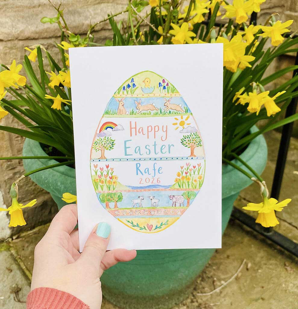 Hand holding a personalised 'Happy Easter' card with an illustrated egg design in front of yellow flowers.