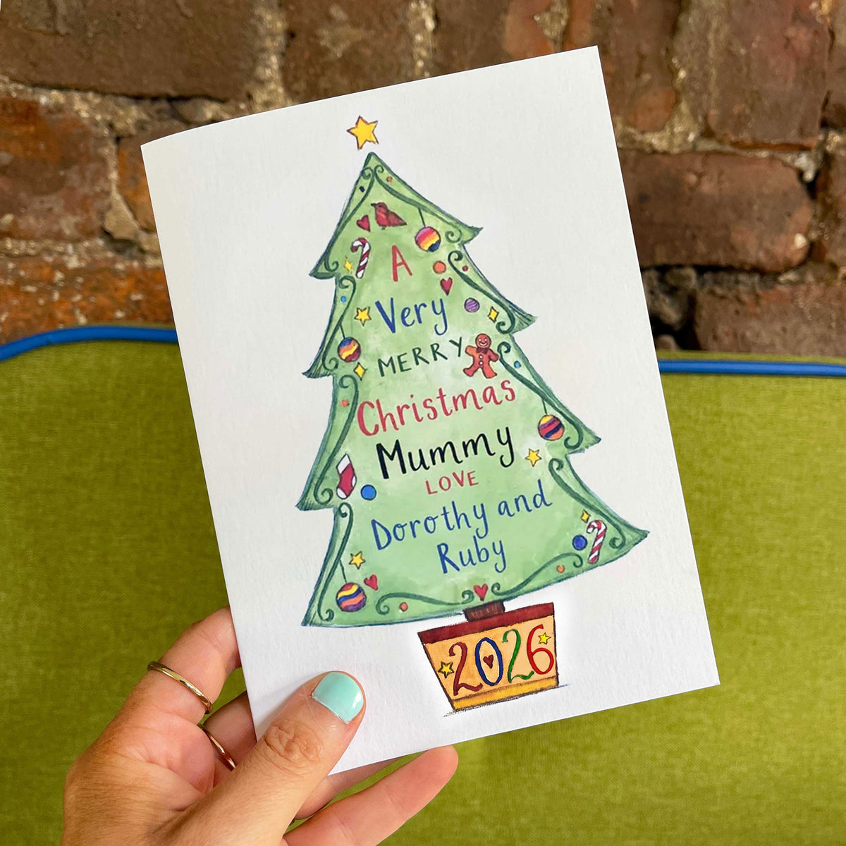 A hand holds a personalised Christmas greeting card featuring a hand-drawn green Christmas tree with the message 'A Very Merry Christmas Mummy Love Dorothy and Ruby' and the year '2026' at the bottom. The background shows a green cushion and a brick wall.