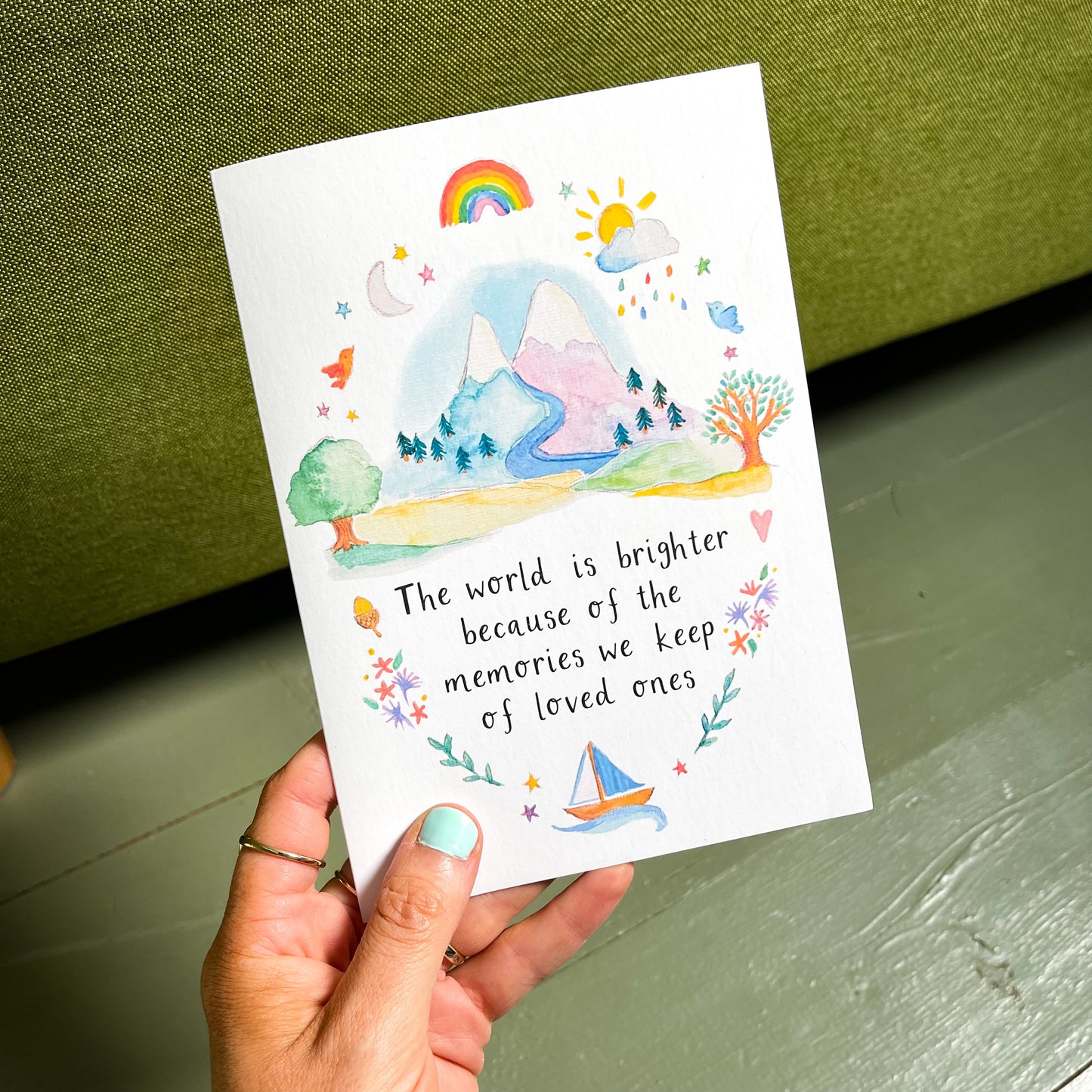 A hand holding a Sympathy Card with a watercolor design featuring a rainbow, sun, tree, and mountain. The text on the card reads: 'The world is brighter because of the memories we keep of loved ones'.