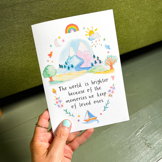 A hand holding a Sympathy Card with a watercolor design featuring a rainbow, sun, tree, and mountain. The text on the card reads: 'The world is brighter because of the memories we keep of loved ones'.
