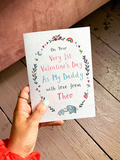 Hand holding a Valentine's Day card for a new dad on a wooden floor.
