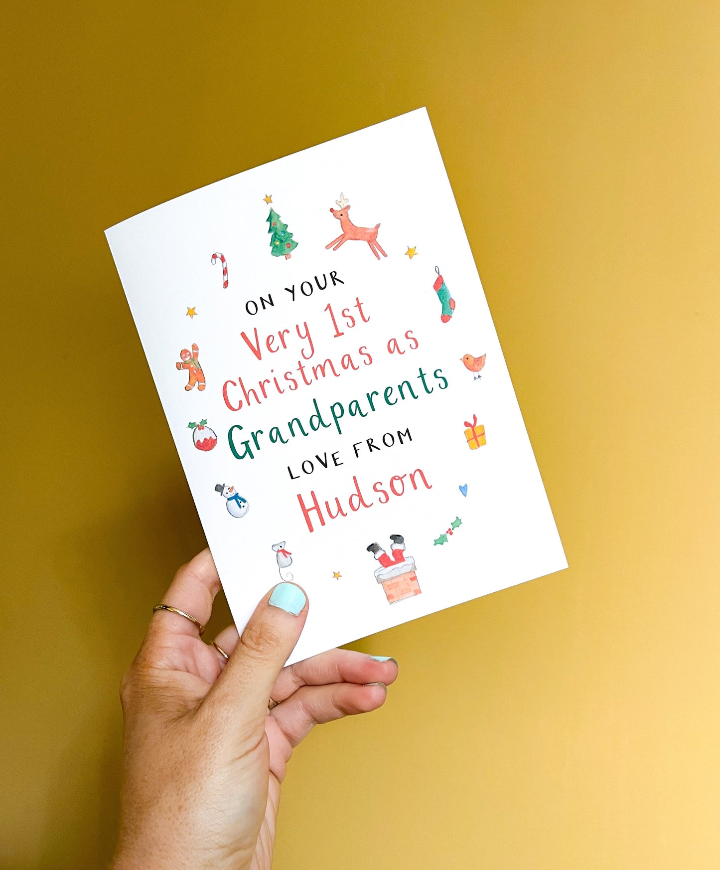 A hand holding a personalised Christmas greetings card for grandparents with illustrations of a Santa hat, reindeer, and Christmas tree. The text on the card reads: 'On your Very 1st Christmas as Grandparents Love From 'Name'.