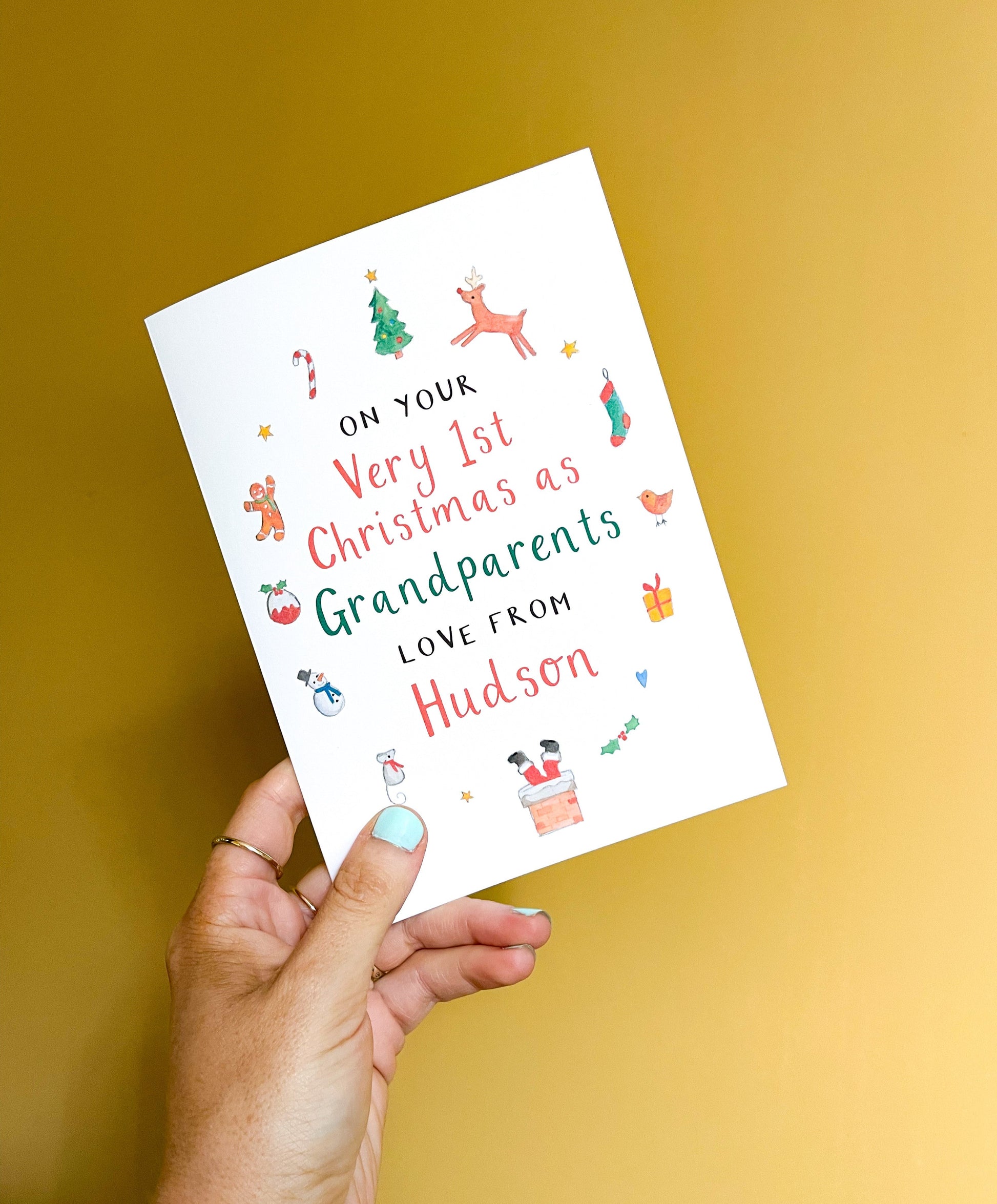 A hand holding a personalised Christmas greetings card for grandparents with illustrations of a Santa hat, reindeer, and Christmas tree. The text on the card reads: 'On your Very 1st Christmas as Grandparents Love From 'Name'.
