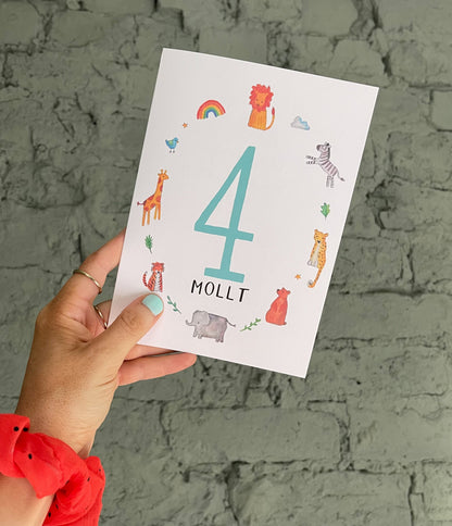 A hand holding a personalised birthday card with the number 4 and the name 'Molly' on it, featuring colorful animal illustrations around the number.