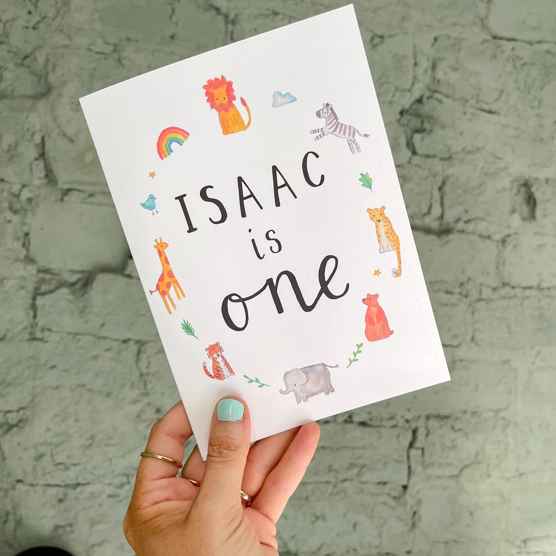 hand holding a white greetings card with watercolour animal illustrations surrounding the personalised text 'isaac is one'