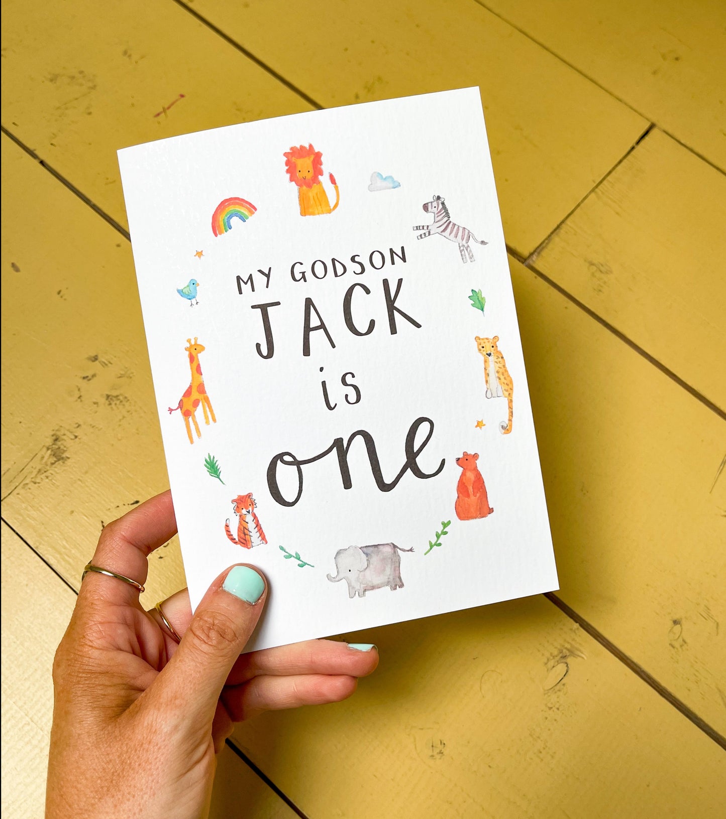 A hand holding a personalised birthday card for a godson with illustrated animals around the text. The card is white with black text and colourful illustrations.