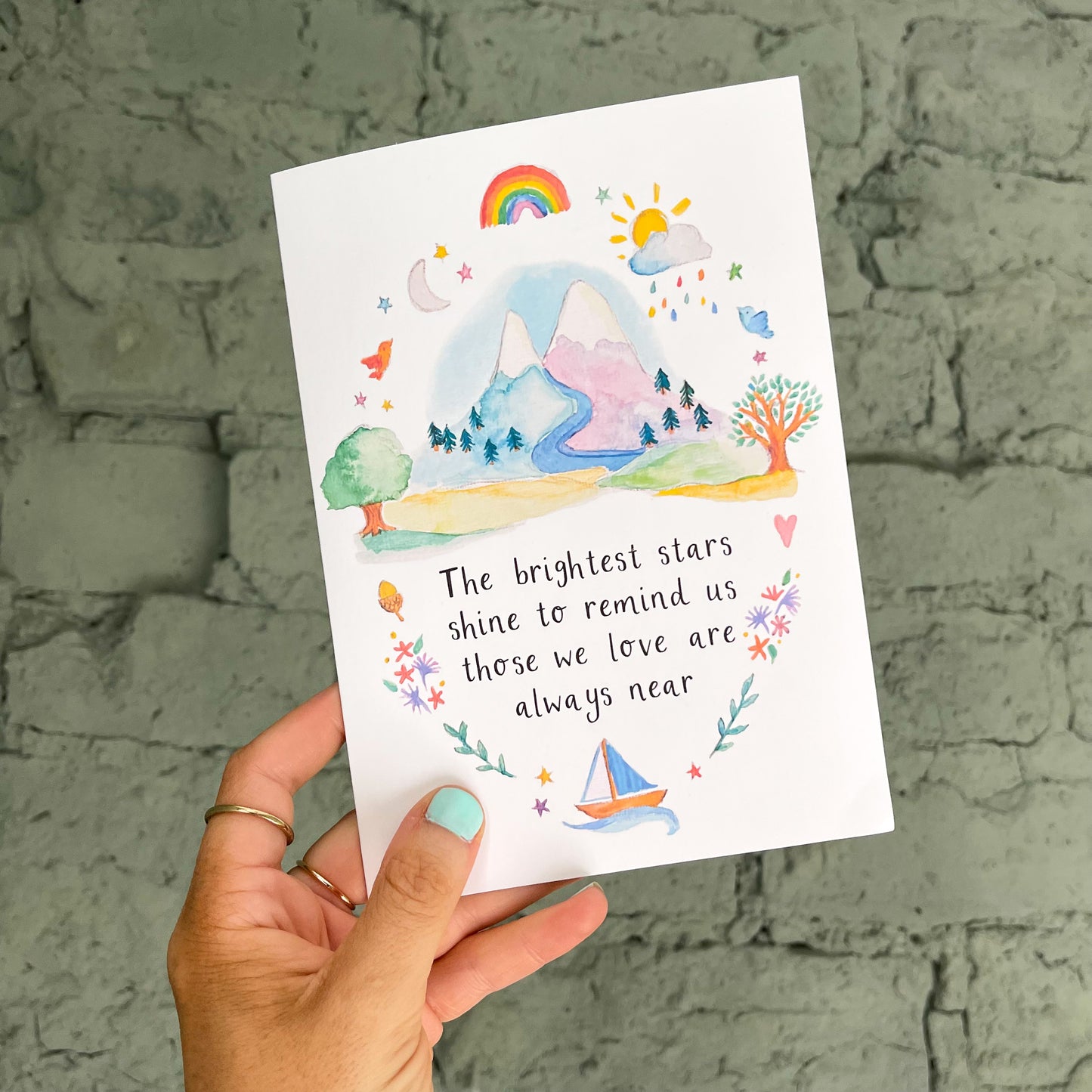 hand holding a white greetings card with mountain watercolour illustration and the text 'the brightest stars shine to remind us those we love are always near'