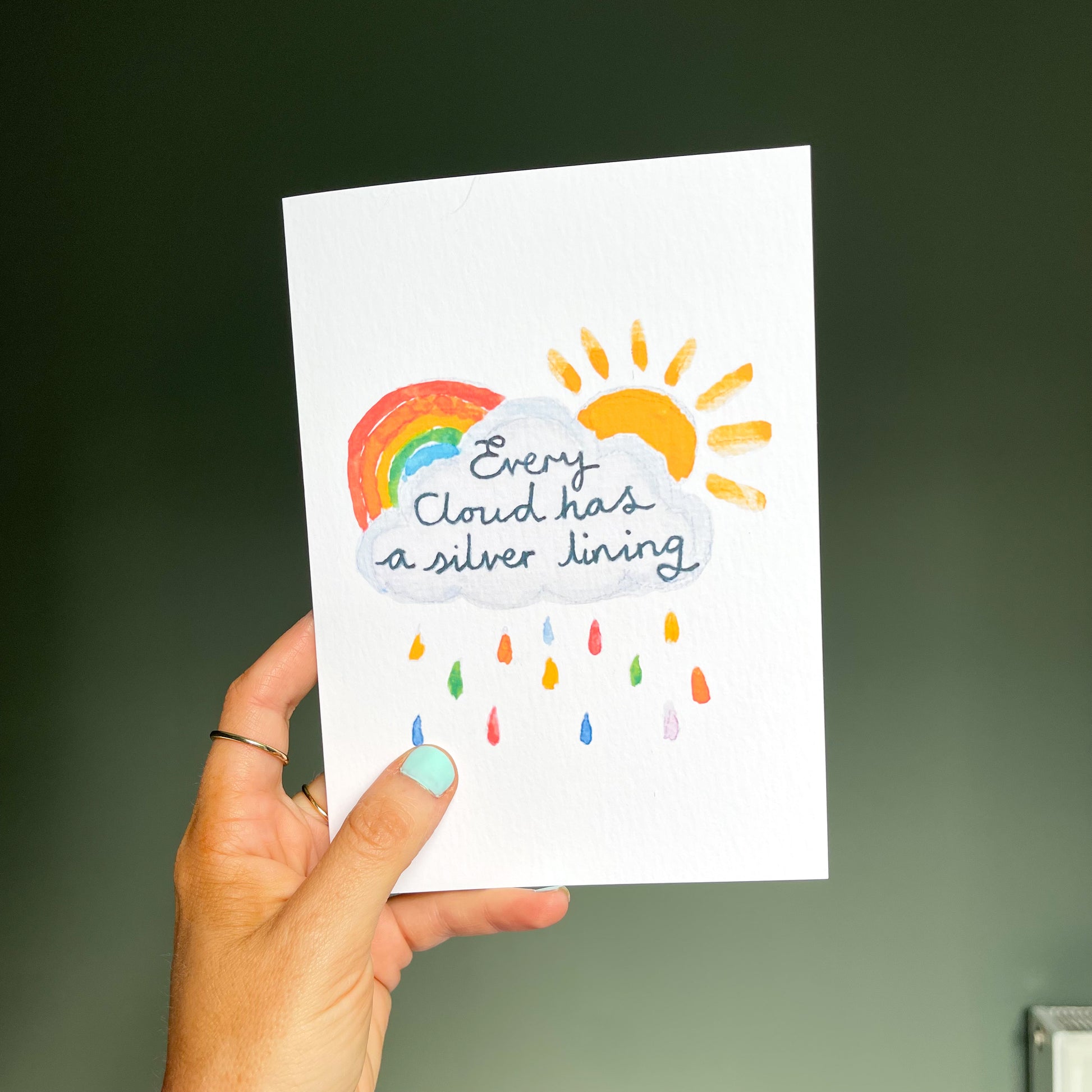 hand holding a white greetings card featuring a watercolour illustration of a rainbow, sun and cloud with the text 'every cloud has a silver lining'