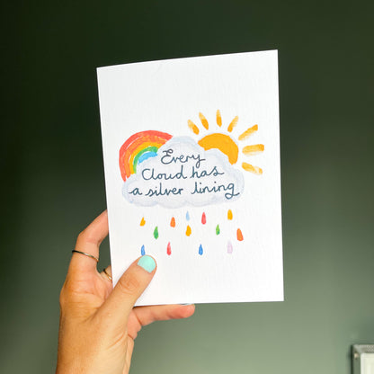 hand holding a white greetings card featuring a watercolour illustration of a rainbow, sun and cloud with the text 'every cloud has a silver lining'