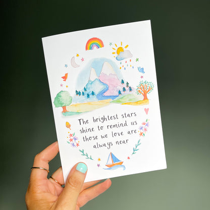 hand holding a white greetings card with mountain watercolour design and the text 'the brightest stars shine to remind us those we love are always near'
