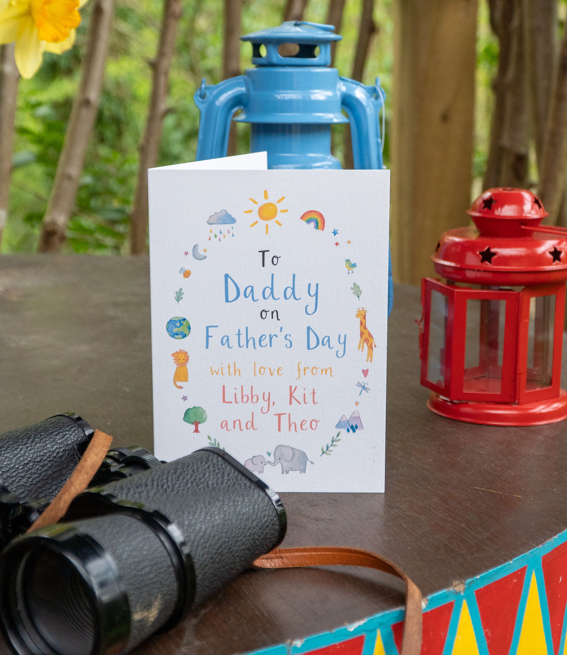 a greetings card with the text 'to daddy on Father's Day with love from libby, kit and theo' stands on a wooden table next to binoculars and a lantern