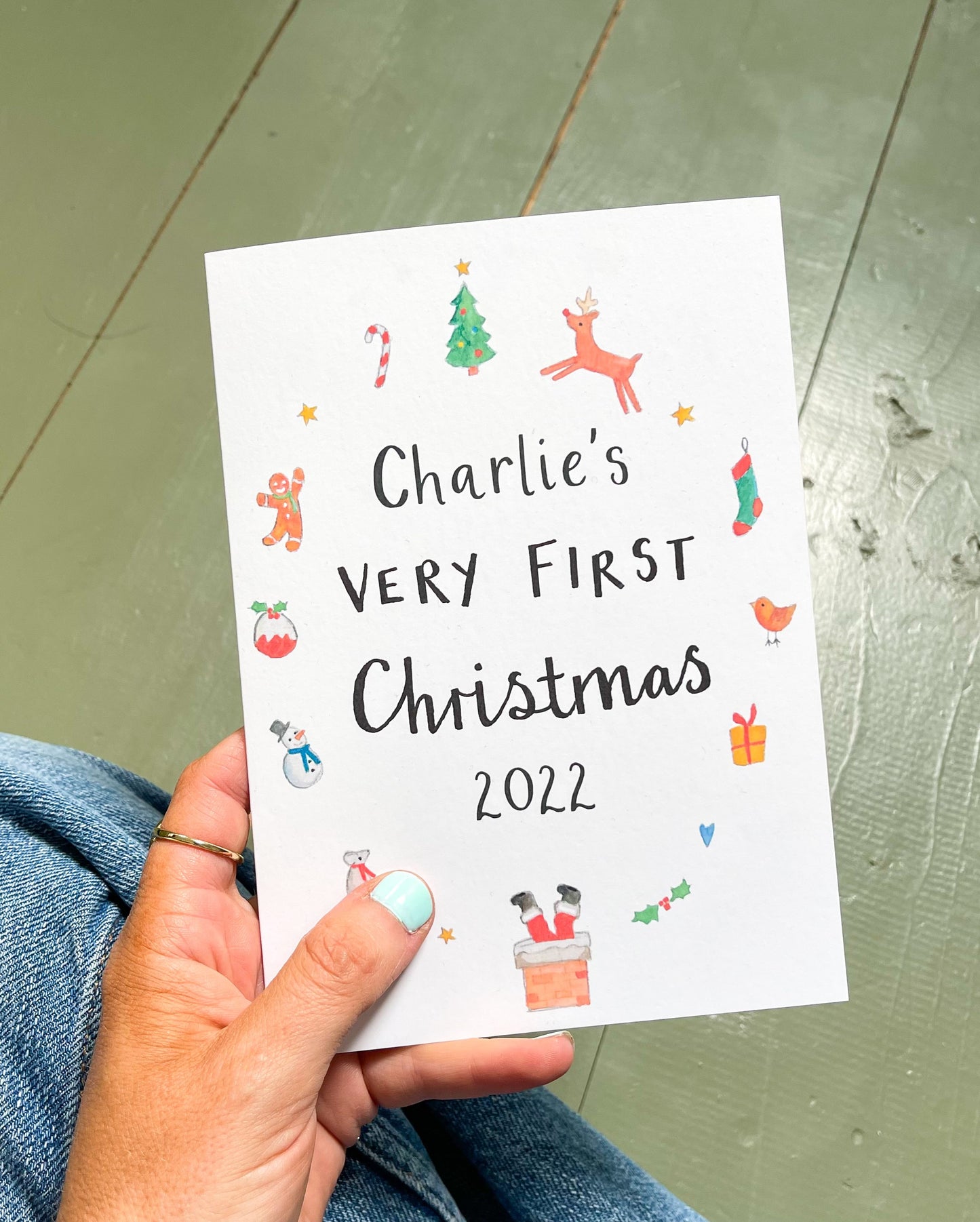 A hand holding a personalised Christmas card with Christmas-themed illustrations and text that reads 'Charlie's Very First Christmas 2022'.